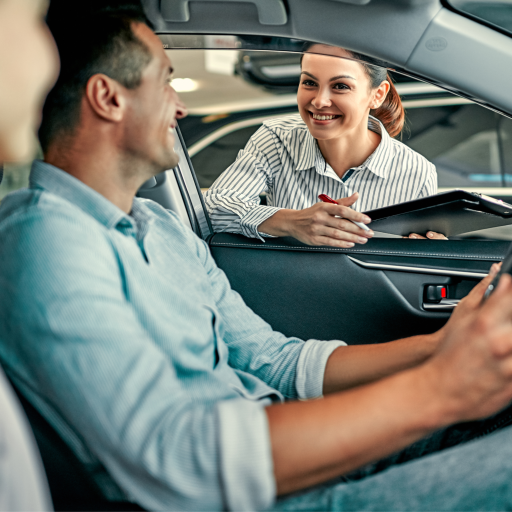 Smiling woman leaning into a car window, talking with a man inside the car.