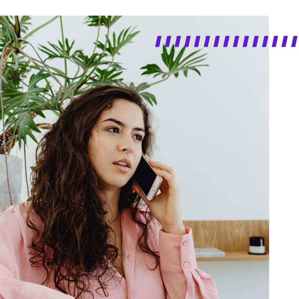 Woman with long curly hair in a pink shirt talks on her phone indoors, with a large leafy plant in the background.